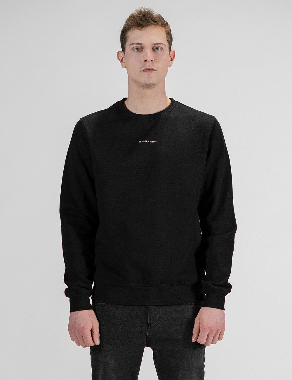 A man with short brown hair stands facing forward against a plain light gray background. He is wearing a long-sleeved, solid black crewneck sweatshirt featuring a small, white text logo centered on the upper chest. He is paired with dark charcoal or black denim pants, which are visible from the waist down. His arms hang relaxed at his sides.