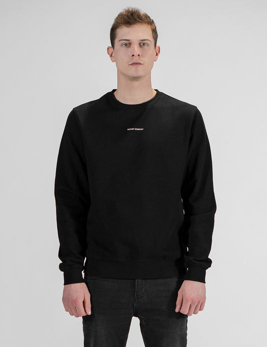 A man with short brown hair stands facing forward against a plain light gray background. He is wearing a long-sleeved, solid black crewneck sweatshirt featuring a small, white text logo centered on the upper chest. He is paired with dark charcoal or black denim pants, which are visible from the waist down. His arms hang relaxed at his sides.