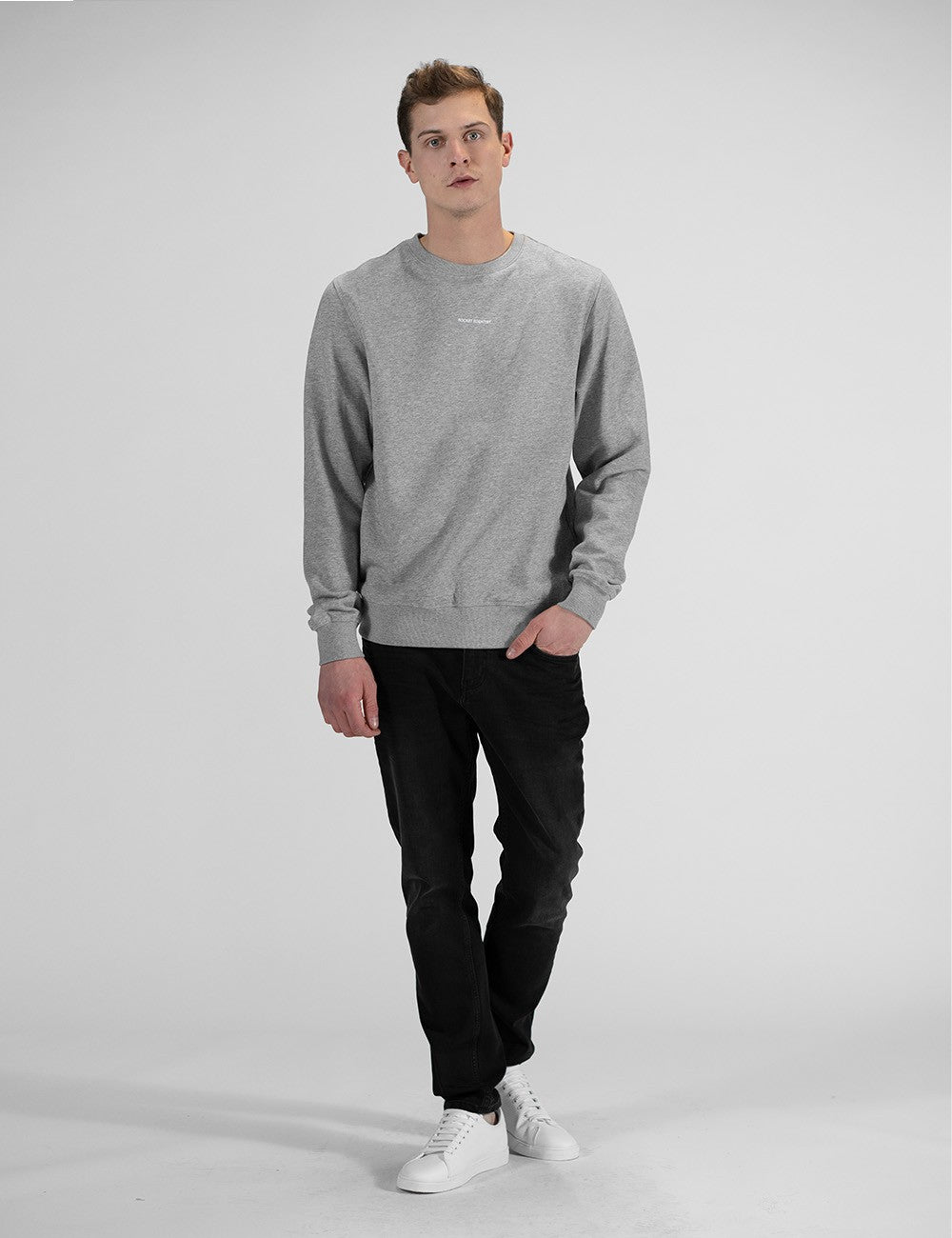 A young man with light brown hair stands against a plain light grey background, wearing a grey Rocket Science crewneck sweatshirt with a small print on the chest, black pants, and white sneakers. 