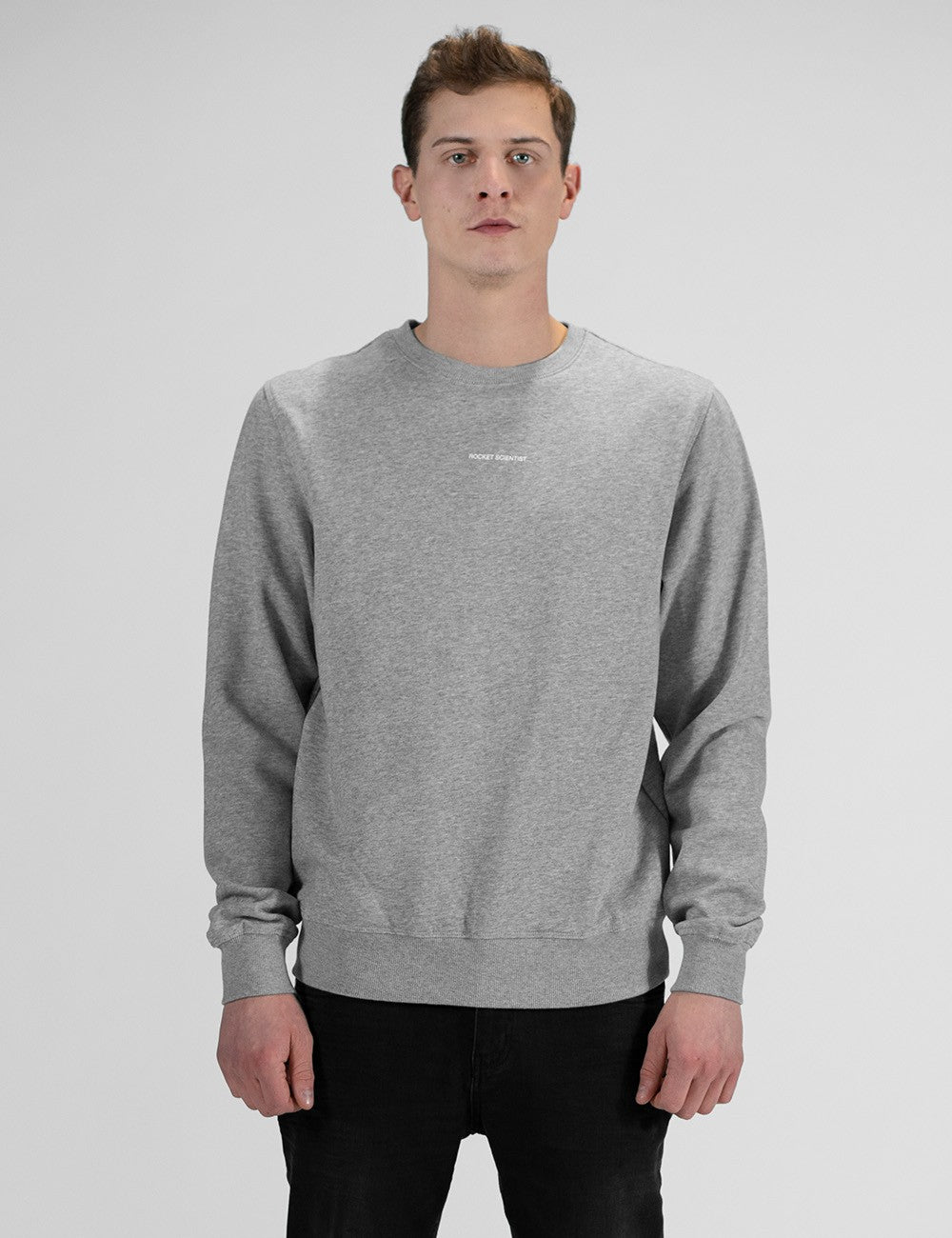 A man with short brown hair in front profile against a plain white background, wearing a heather grey crewneck sweatshirt with a small Rocket Scientist print and black pants.