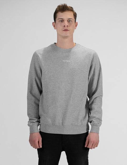 A man with short brown hair in front profile against a plain white background, wearing a heather grey crewneck sweatshirt with a small Rocket Scientist print and black pants.