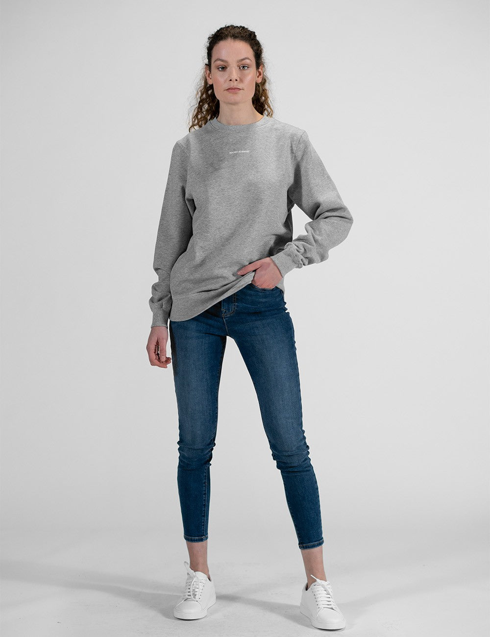 A young woman stands facing forward against a plain light grey background, wearing a grey crew neck sweatshirt, dark blue skinny jeans, and white sneakers.
