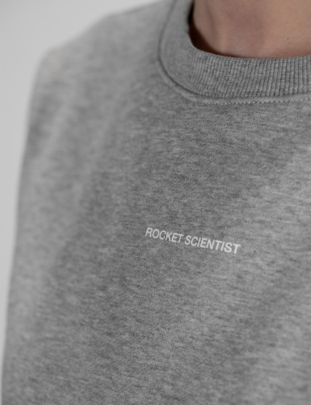 A close-up of a person wearing a heather grey crewneck sweatshirt. The fabric has a classic textured, speckled grey appearance. Small, white, sans-serif text reading "ROCKET SCIENTIST" is printed or embroidered horizontally across the left chest area. The ribbed neckline of the sweatshirt is visible at the top right, fitting closely around the person's neck. The background is a clean, neutral, light grey, and the shot is taken from a slightly angled, side-profile perspective.