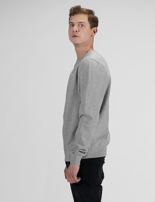 A man with short brown hair stands in profile against a plain white background, wearing a heather grey crewneck sweatshirt and black pants.