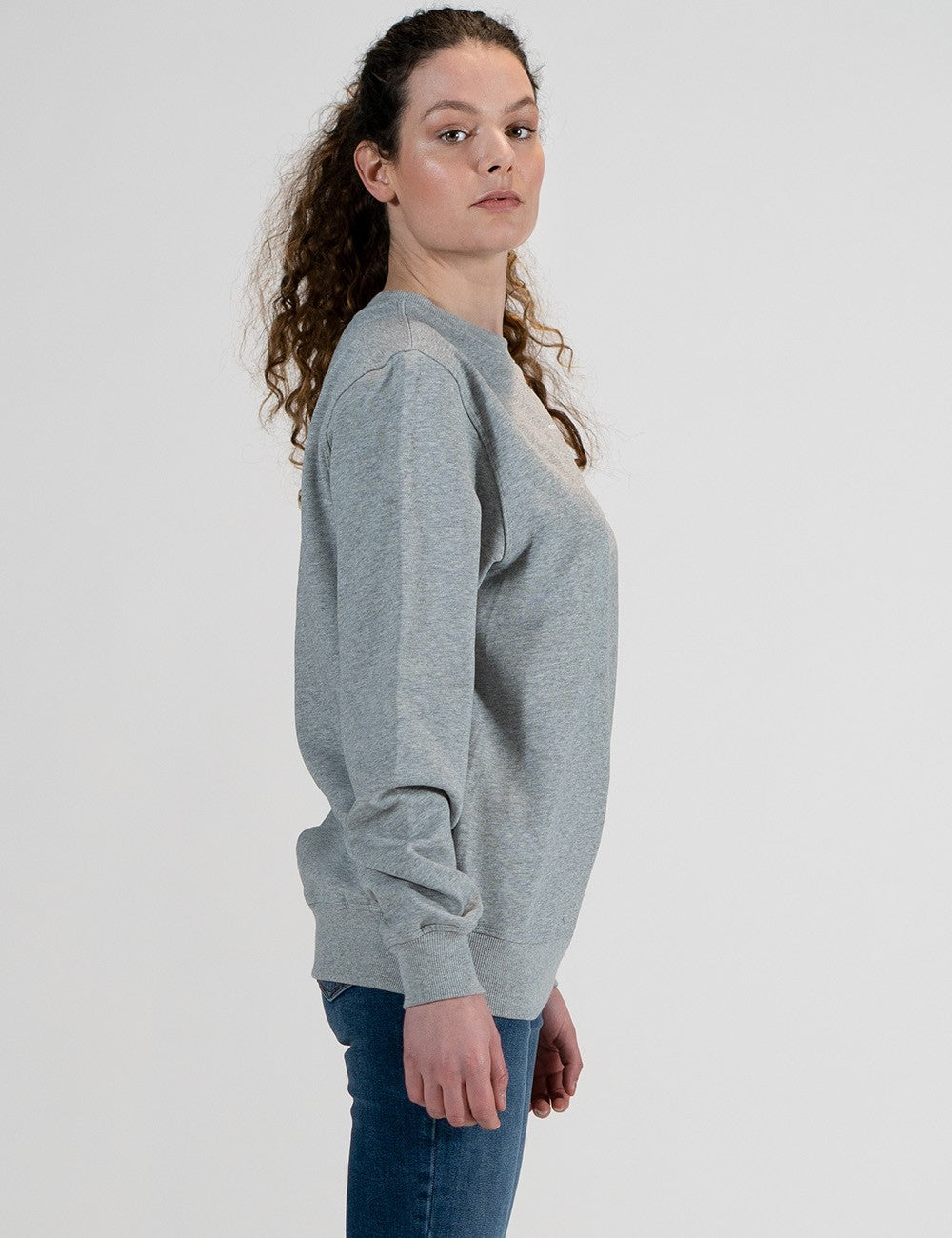 A woman with curly brown hair tied back stands in profile against a plain, light grey background. She is wearing a classic grey crewneck sweatshirt with long sleeves and ribbed cuffs, paired with blue jeans.
