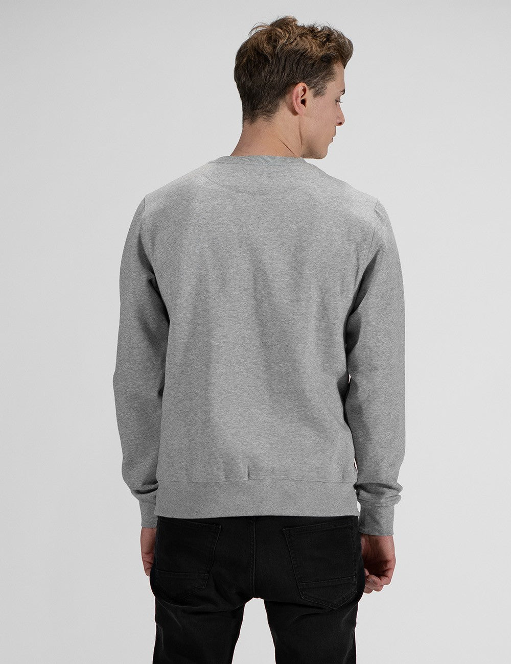 A young man with light brown, styled wavy hair is shown from the back, looking slightly to his right. He is wearing a light grey crew-neck sweatshirt with ribbed cuffs and a ribbed waistband, paired with black pants. The background is a solid, neutral light grey.