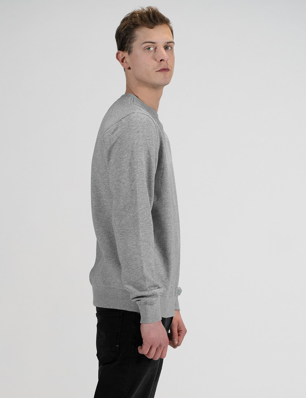 A young man with short brown hair stands in profile, facing right and looking back over his shoulder toward the camera against a plain, light grey background. He is wearing a classic grey marl crewneck sweatshirt with ribbed cuffs and a ribbed waistband, paired with black pants.
