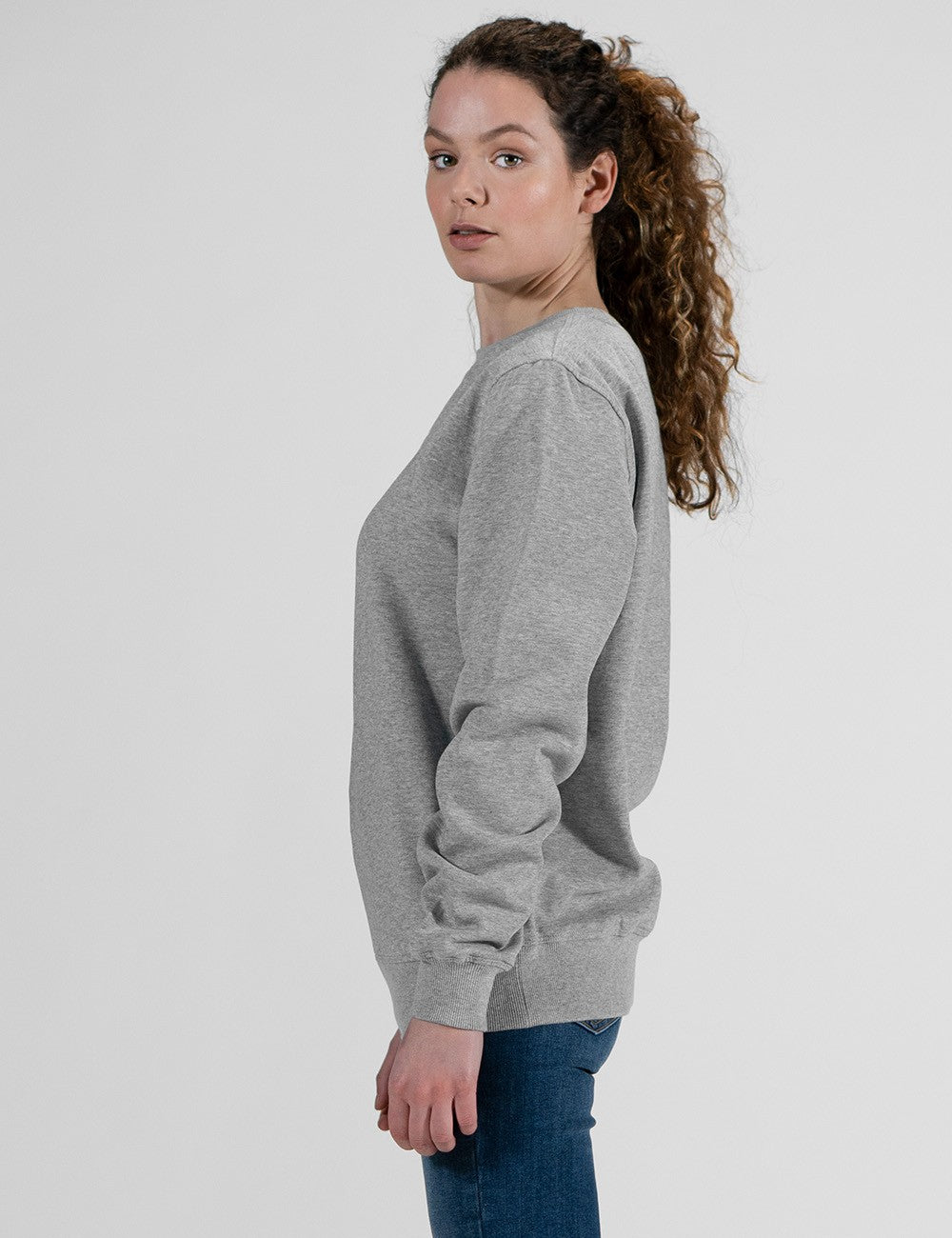 A side-profile shot of a woman with long, curly brown hair tied in a ponytail, wearing a light gray crewneck sweatshirt and blue jeans against a plain white background.