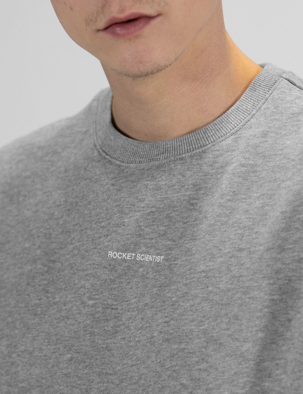 A close-up shot of a person wearing a grey marl crewneck sweatshirt. The focus is on the upper chest and collar area, highlighting a small, white, minimalist text print that reads "ROCKET SCIENTIST" centered on the chest.