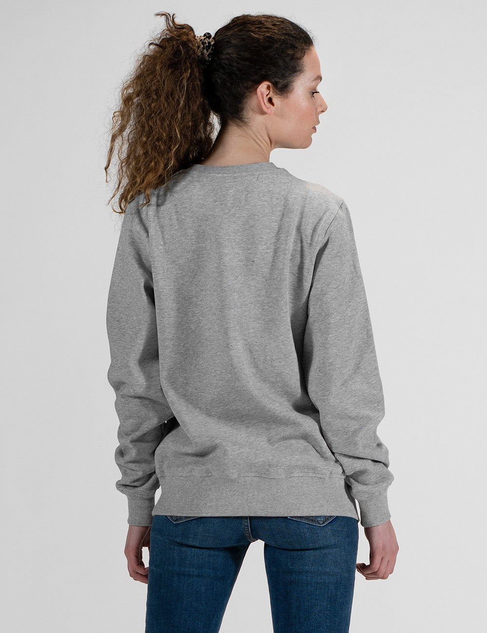 A woman with curly brown hair tied in a ponytail is shown from behind wearing a grey crewneck sweatshirt and blue jeans.