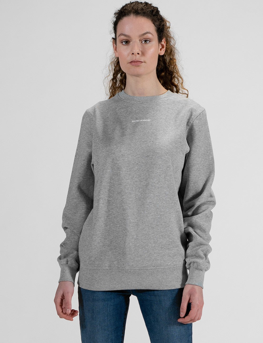 A woman with curly brown hair tied back stands against a plain light grey background, wearing a heather grey crewneck sweatshirt and blue jeans. The long-sleeved sweatshirt has a relaxed fit with ribbed cuffs and a ribbed hem, and features a small, subtle white logo printed horizontally across the center of the upper chest.