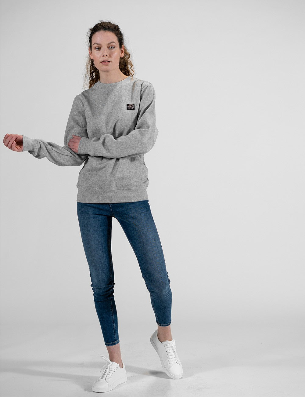 A woman modeling a light gray crewneck sweatshirt, blue skinny jeans, and white sneakers against a plain light gray studio background.