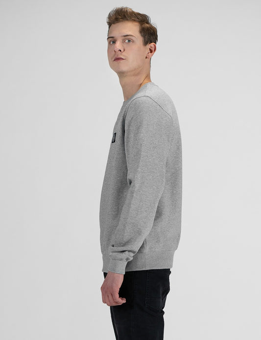 A man wearing a light gray heather crewneck sweatshirt with a small black chest logo, shown in a side profile against a light gray studio background.