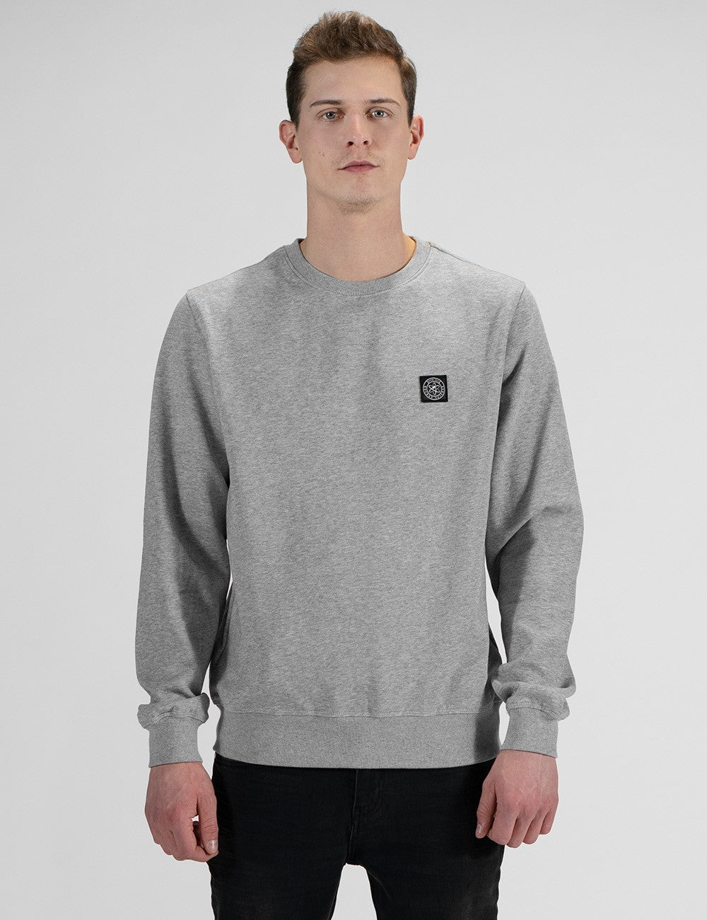 A man wearing a light gray heather crewneck sweatshirt with a small black square logo patch on the left chest, paired with black pants, against a light gray studio background.