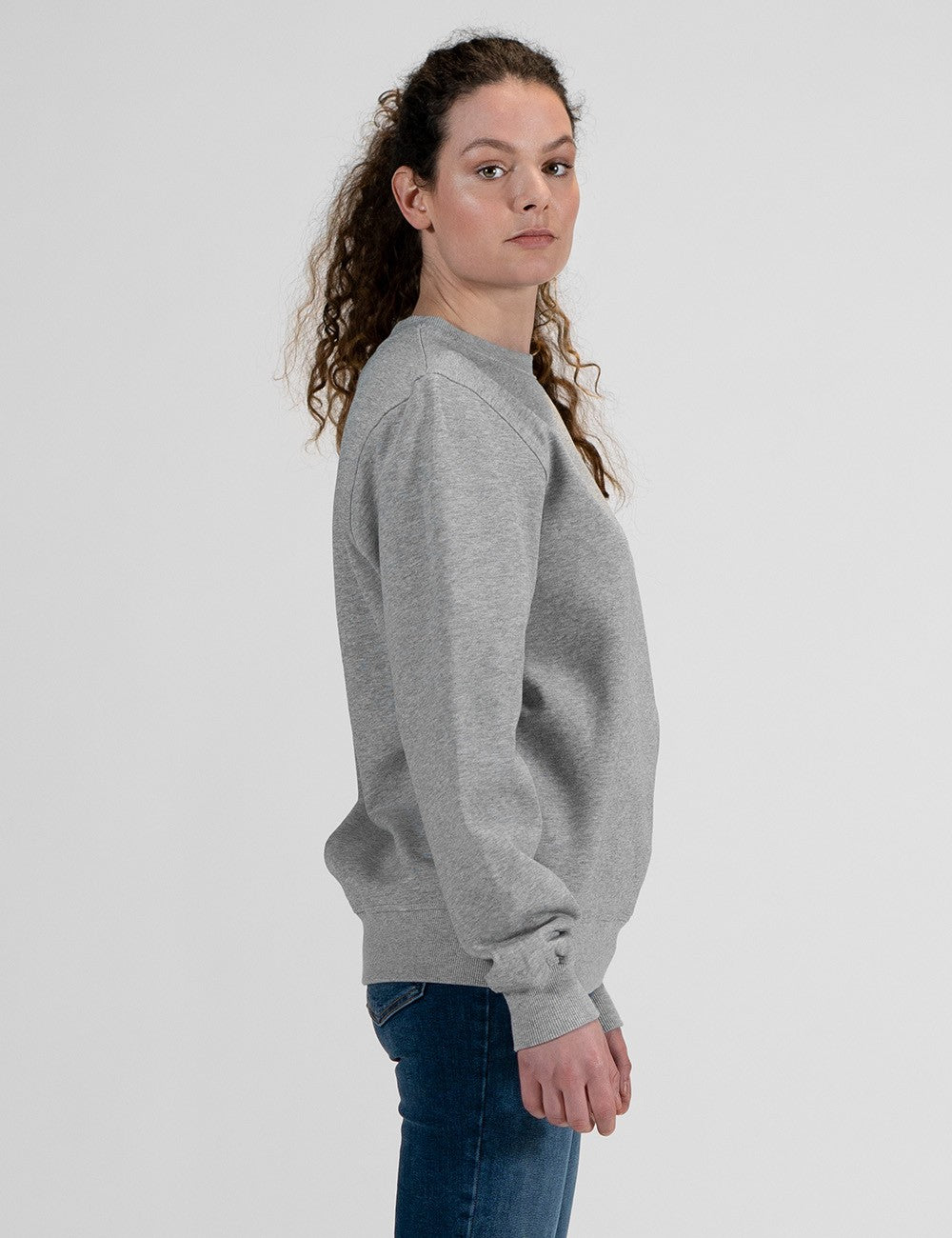 A woman shown in side profile wearing a light gray crewneck sweatshirt and blue jeans against a plain light gray studio background.