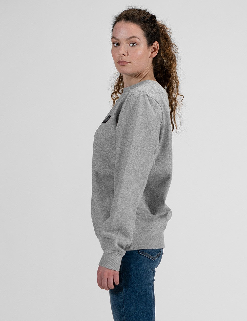 A woman wearing a light gray crewneck sweatshirt and blue jeans, shown from a side profile against a plain light gray studio background.