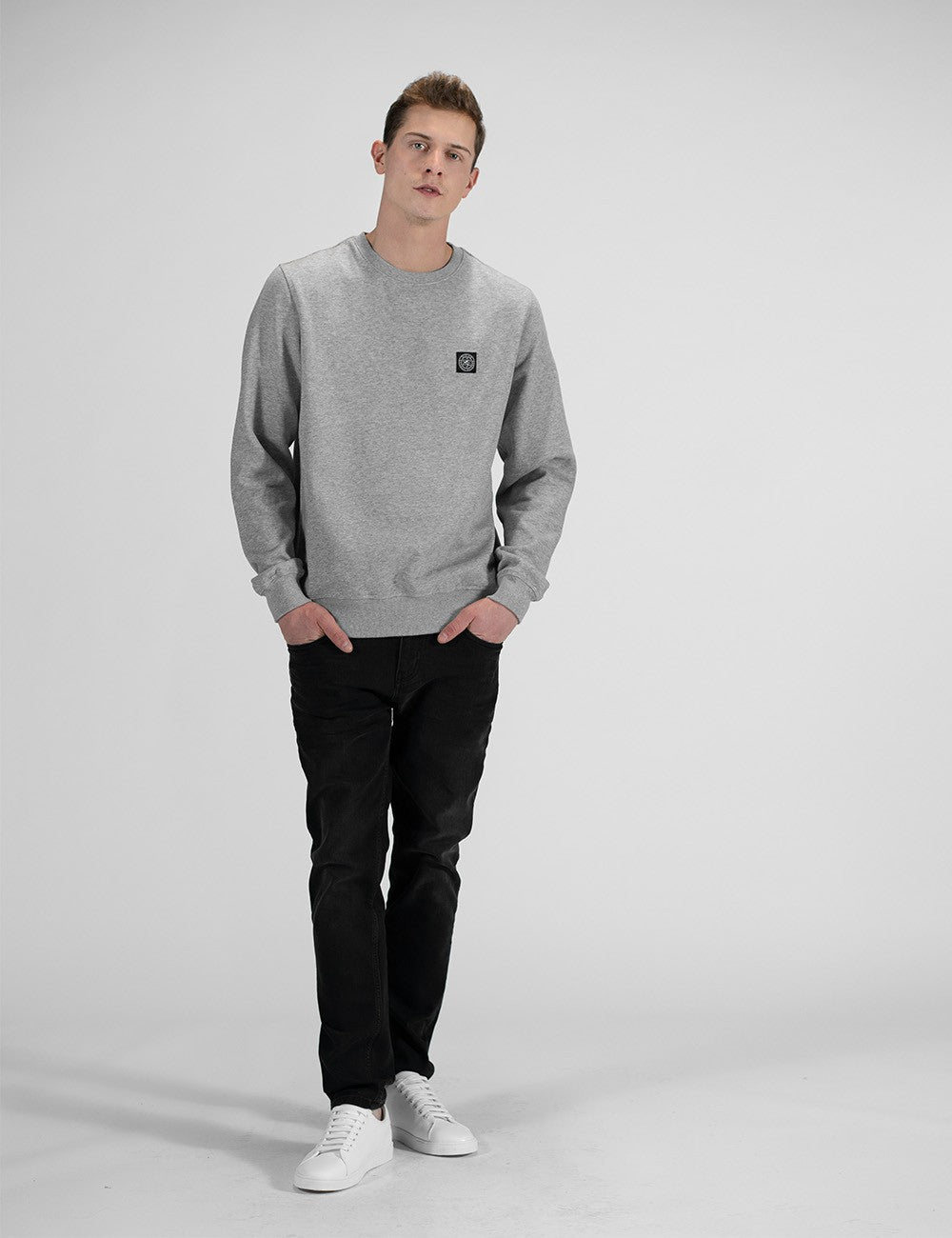 A man modeling a light gray crewneck sweatshirt with a small black square logo on the left chest, paired with black pants and white sneakers, standing against a light gray studio background.