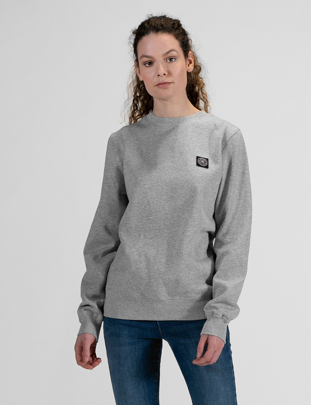 A woman modeling a light gray crewneck sweatshirt with a small black rectangular logo patch on the left chest, paired with blue jeans, against a solid light gray background.