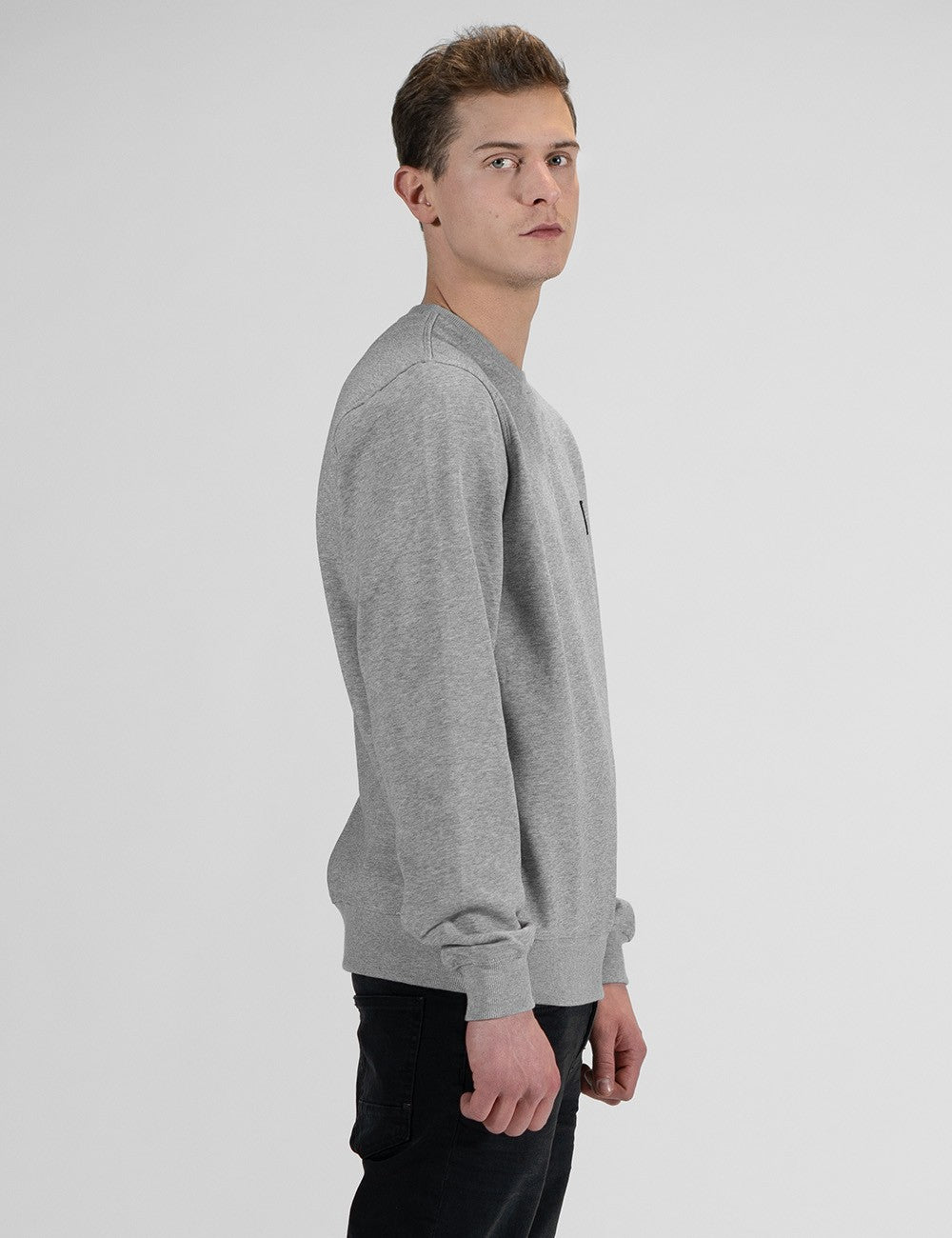 A man in a side profile wearing a light gray crewneck sweatshirt and black pants, photographed against a neutral light gray studio background.