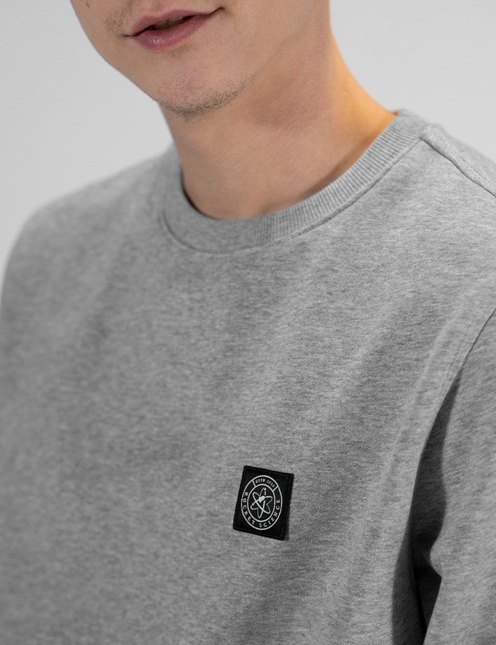 Close-up of a person wearing a heather gray crewneck sweatshirt with a black square chest patch featuring a white circular logo.