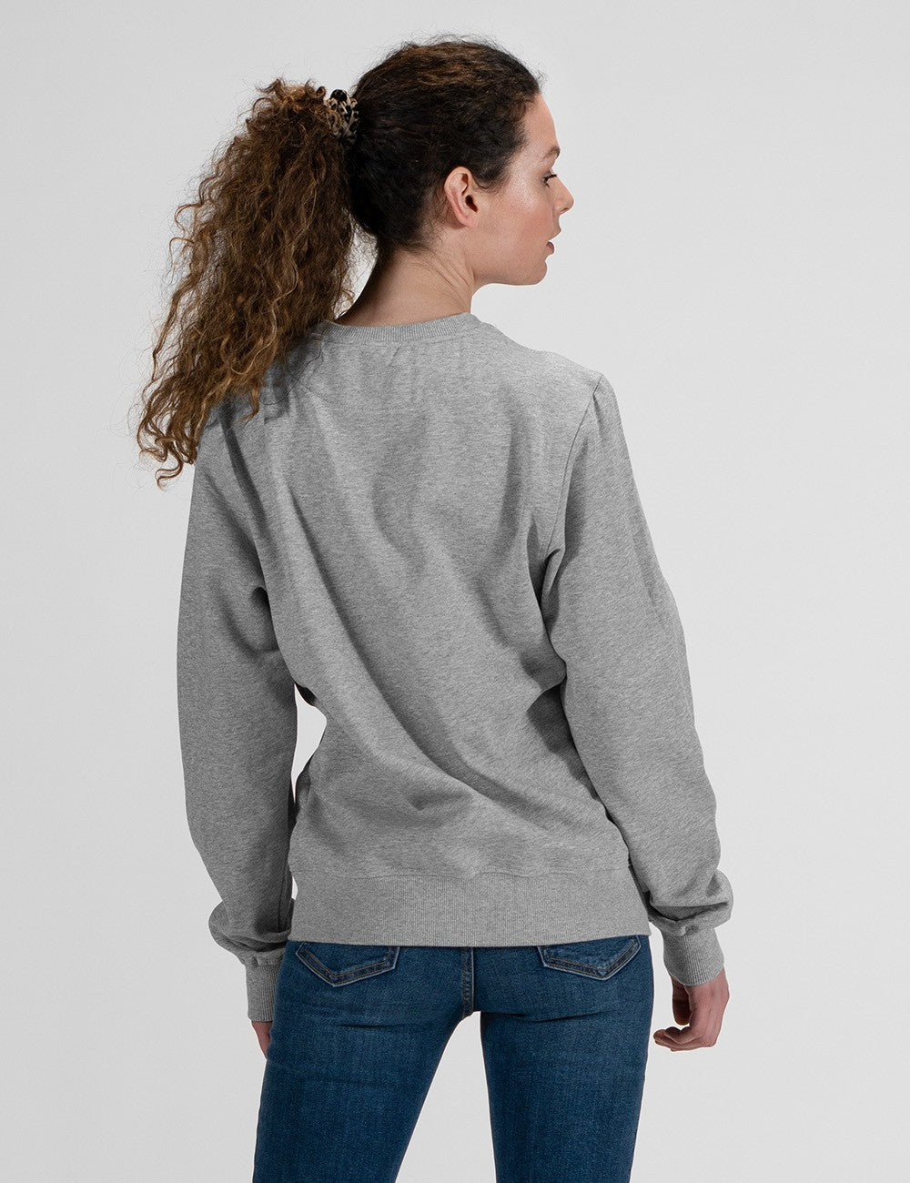 A woman with curly brown hair in a ponytail, viewed from the back, wearing a light gray crewneck sweatshirt and blue jeans against a light gray studio background.