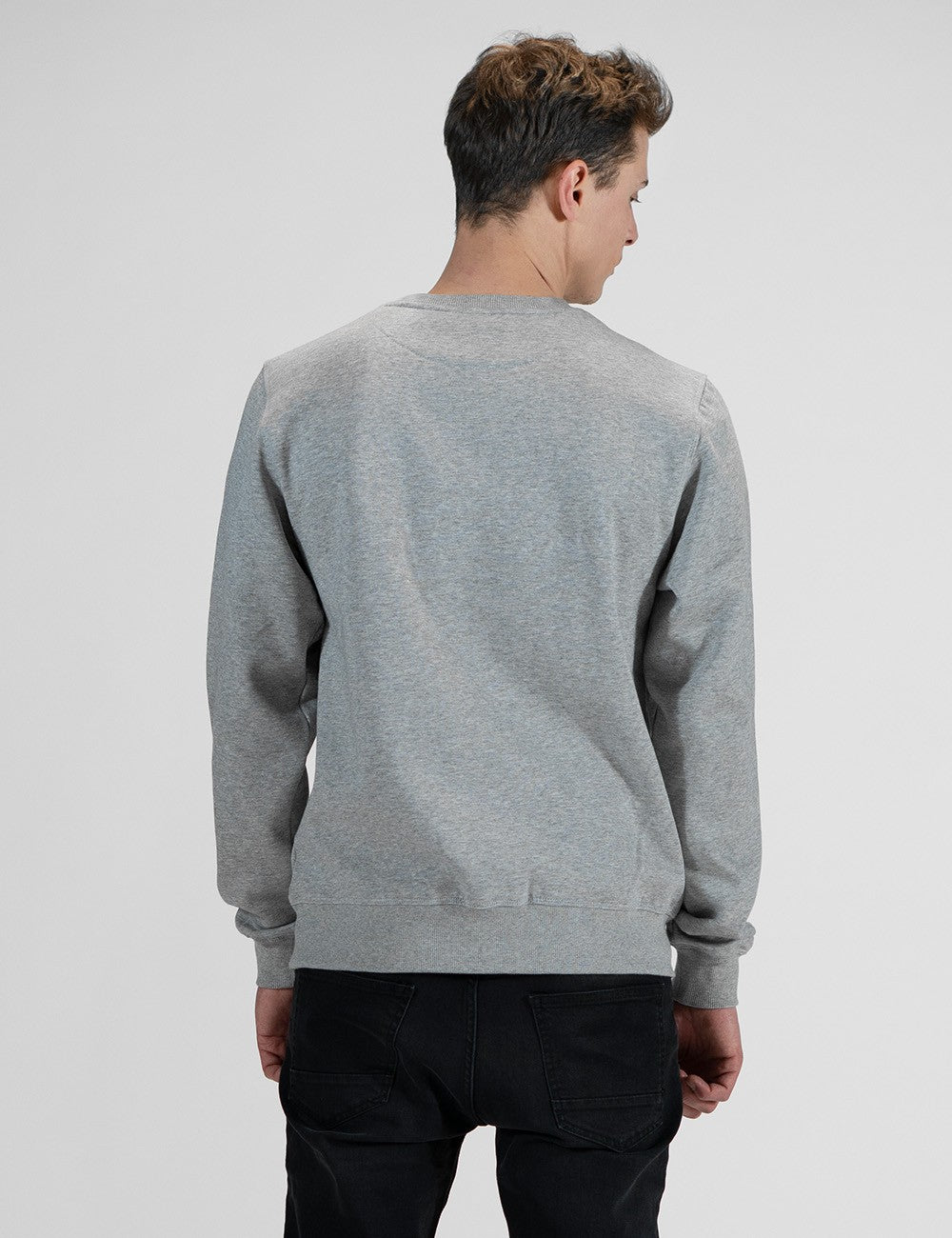 A man viewed from behind wearing a light gray heather crewneck sweatshirt and black pants, standing against a light gray studio background.