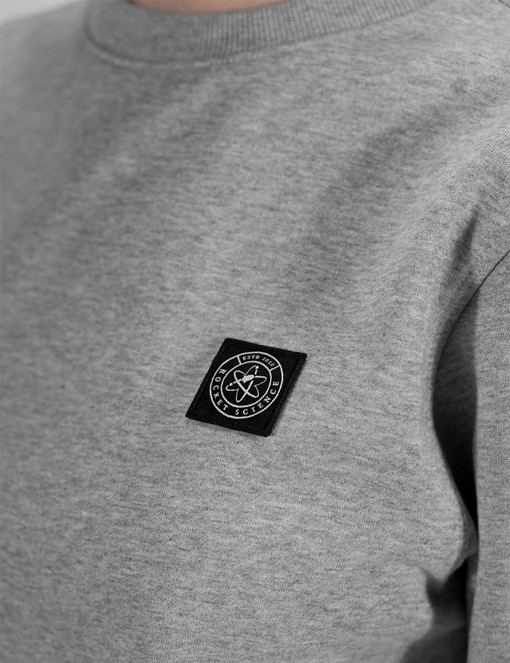 Close-up of a heather gray crewneck sweatshirt featuring a black square chest patch with a white circular logo that reads 'Rocket Science'.