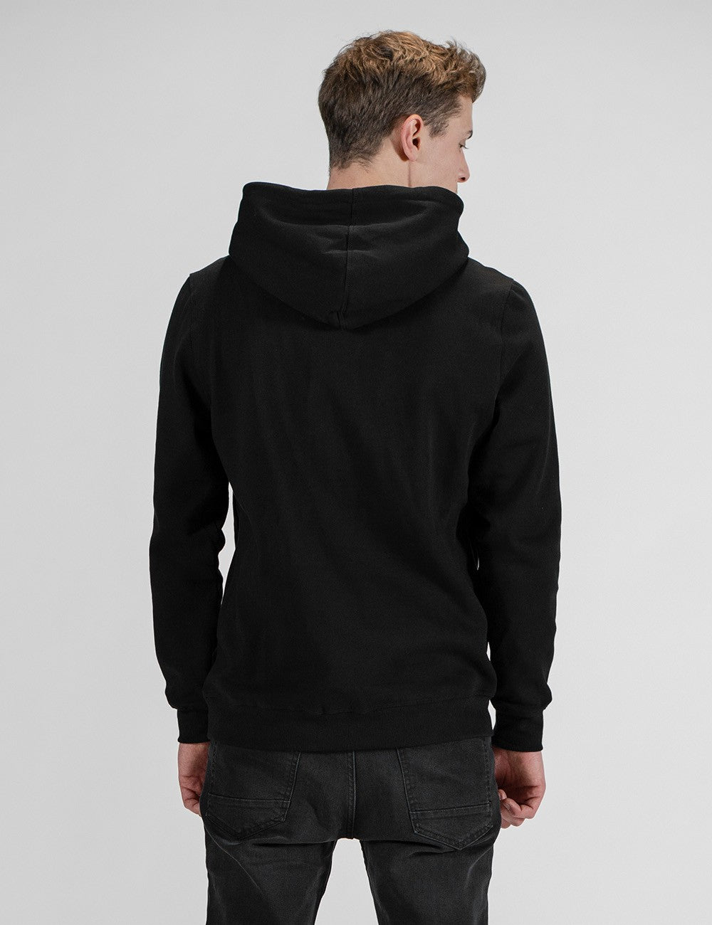 A back view of a man with short blonde hair wearing a solid black pullover hoodie and dark pants, standing against a light grey background while looking slightly to his right.