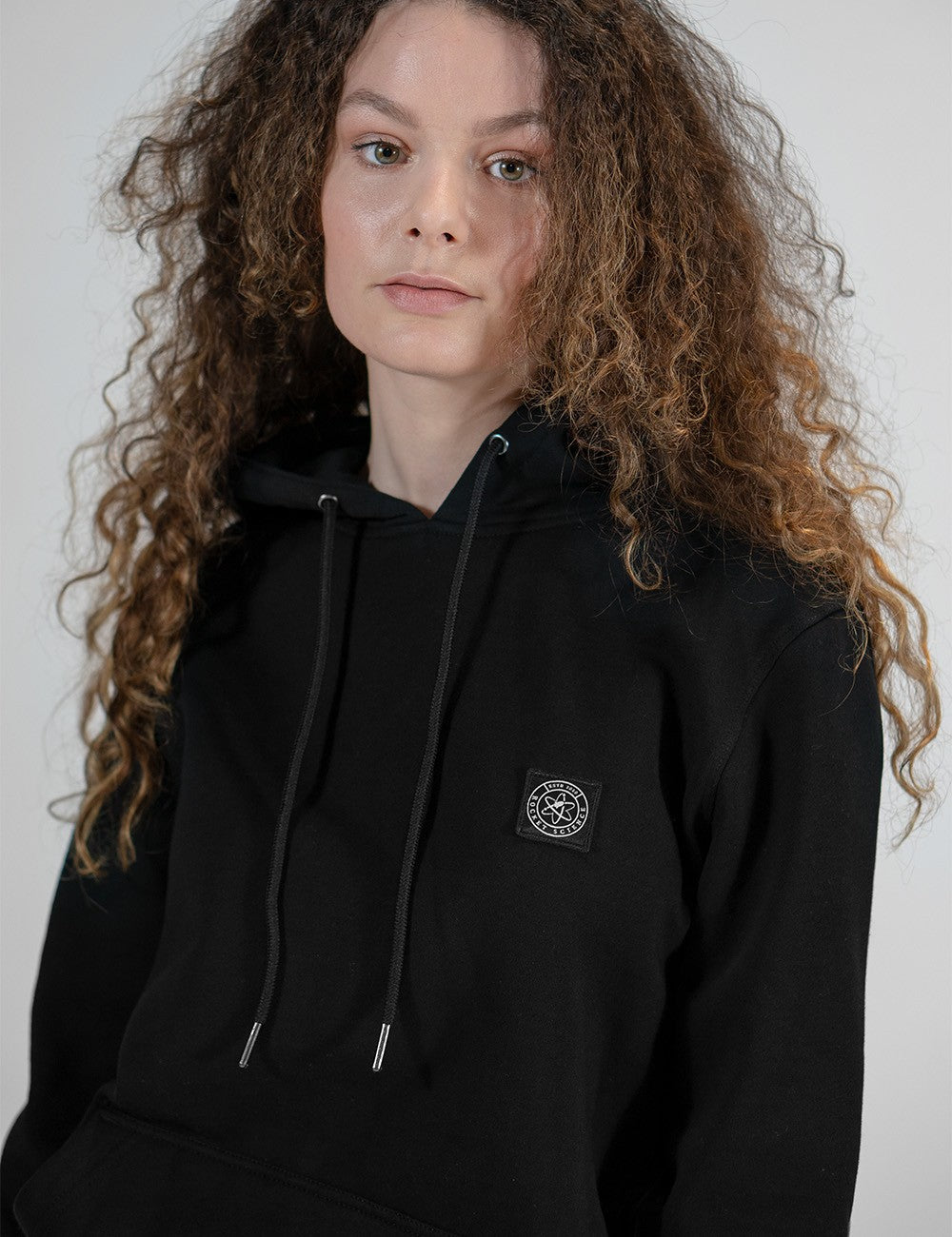 A portrait of a woman with voluminous, curly, shoulder-length brown hair, wearing a solid black pullover hoodie. The hoodie features matching black drawstrings with metallic silver tips and a small, circular black-and-white patch on the left side of the chest. 