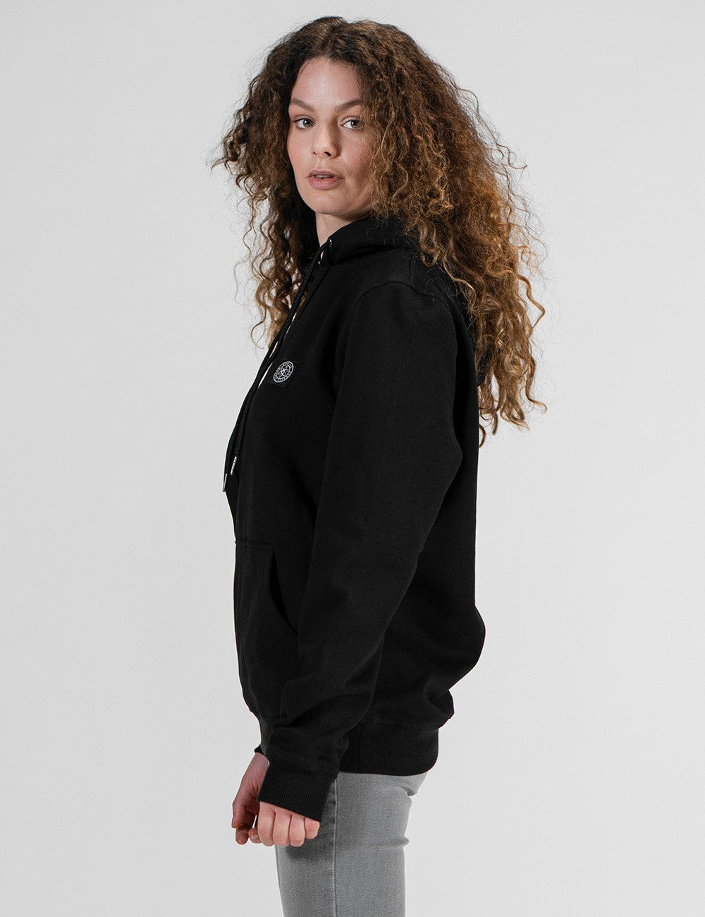A woman with long, curly blonde and brown hair, showcasing a black  hoodie and light gray jeans against a plain white background.