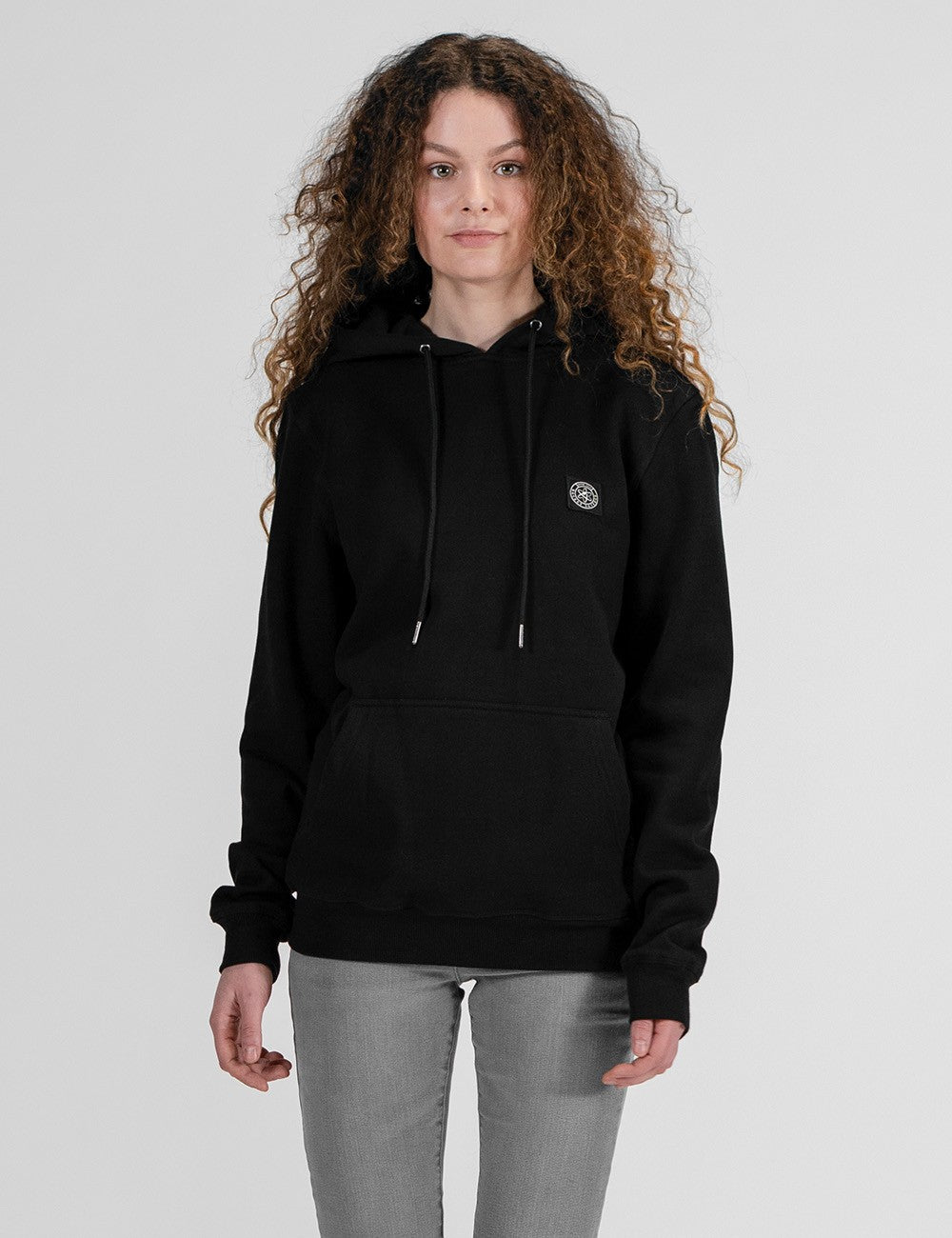 A woman with voluminous, long curly brown hair poses against a plain, light grey background. She is wearing a black hooded sweatshirt featuring long sleeves, matching black drawstrings, a front kangaroo pocket, and a small, circular logo patch on the left side of the chest. She is also wearing fitted, light grey jeans.