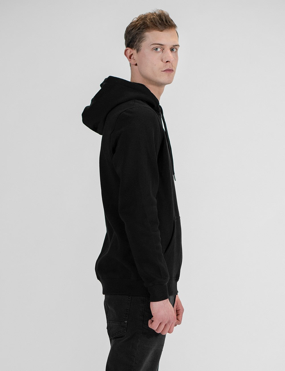 A side-profile shot of a young man with short brown hair looking over his shoulder toward the camera against a plain gray background. He is wearing a plain black pullover hoodie with the hood up slightly at the back, paired with black jeans.