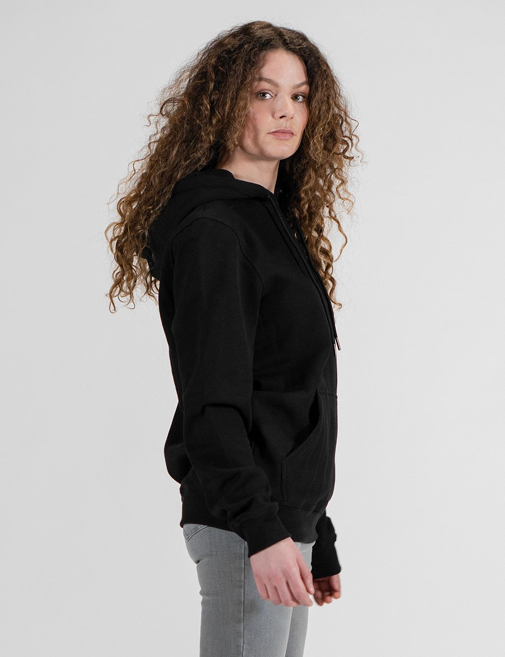 A side view of a person with long, voluminous, curly brown hair wearing a solid black zip-up hoodie and light grey pants, posing against a light grey background.
