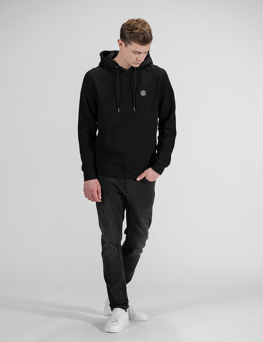 A young man with light brown hair is walking forward against a plain, light grey background, looking down toward his left. He is wearing a black pullover hoodie with drawstrings and a small circular logo on the left chest, paired with slim-fit dark grey or faded black jeans and clean white sneakers. His right hand is casually tucked into his jeans pocket.