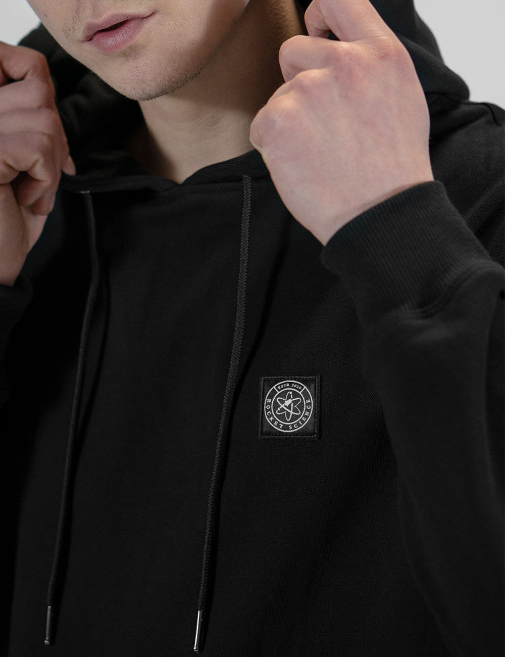 A close-up shot of a person wearing a black pullover hoodie. The individual is holding the drawstrings near the collar with both hands, partially obscuring the neck. On the left breast of the hoodie, there is a small, square black patch containing a white circular logo that features an atom symbol in the center. The background is neutral and slightly out of focus.