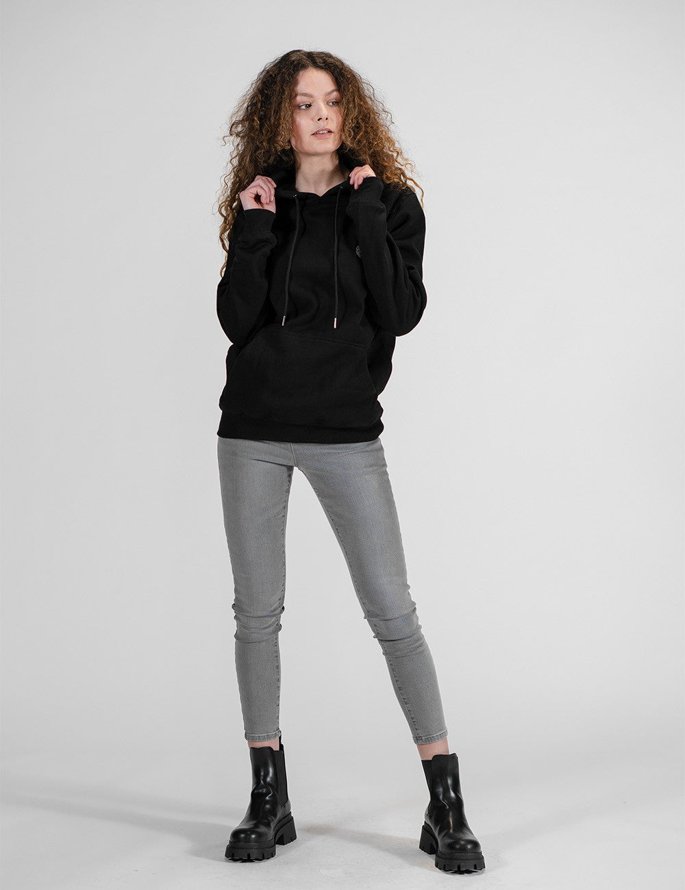 A portrait of a woman with voluminous, curly, shoulder-length brown hair, wearing a solid black pullover hoodie. The hoodie features matching black drawstrings with metallic silver tips and a small, circular black-and-white patch on the left side of the chest. 