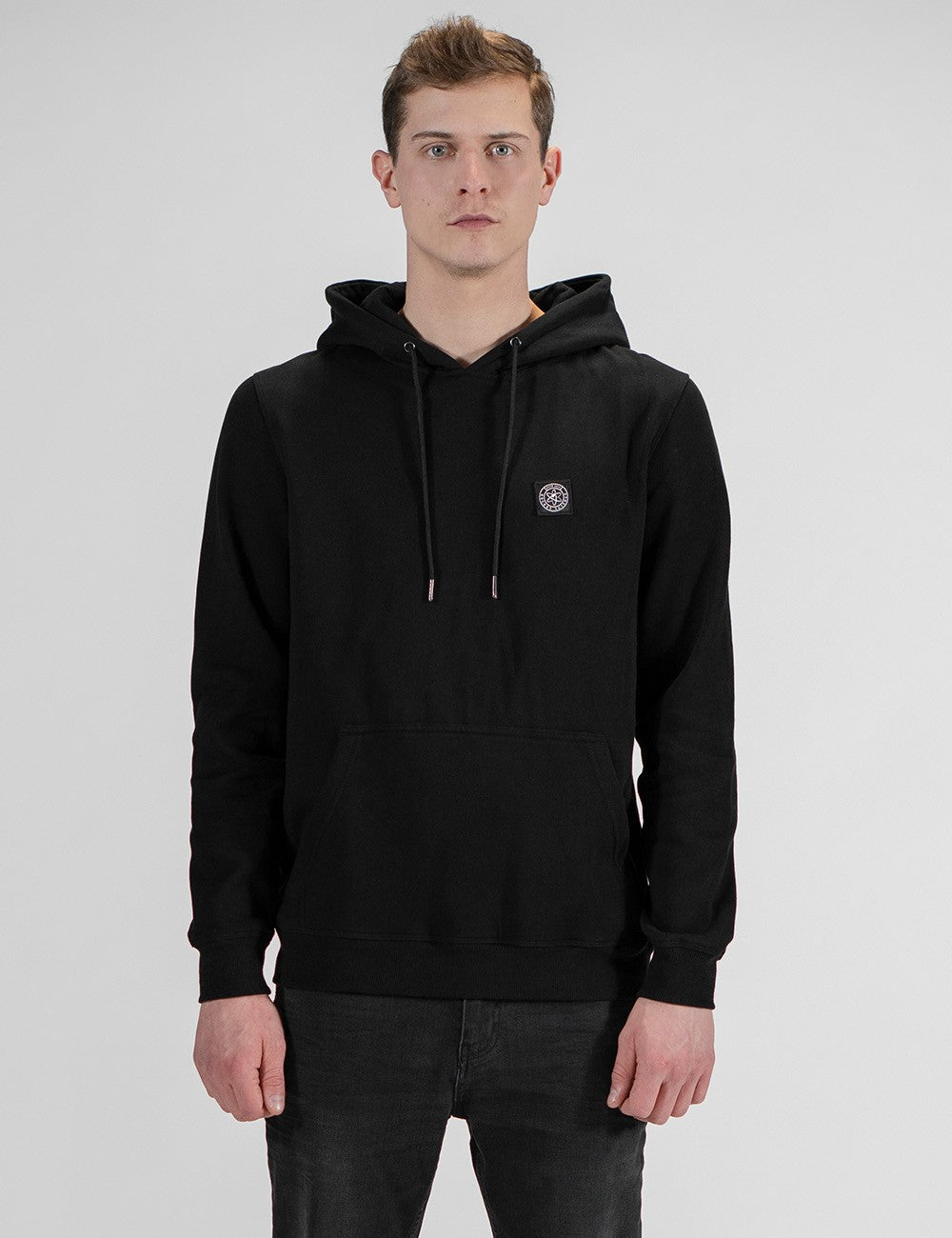 A front-facing, waist-up portrait of a fair-skinned young man with short brown hair, wearing a black drawstring hoodie with a small circular logo patch on the left chest, and dark jeans against a plain light gray background.