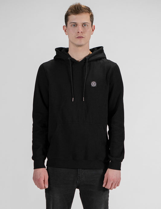 A front-facing, waist-up portrait of a fair-skinned young man with short brown hair, wearing a black drawstring hoodie with a small circular logo patch on the left chest, and dark jeans against a plain light gray background.