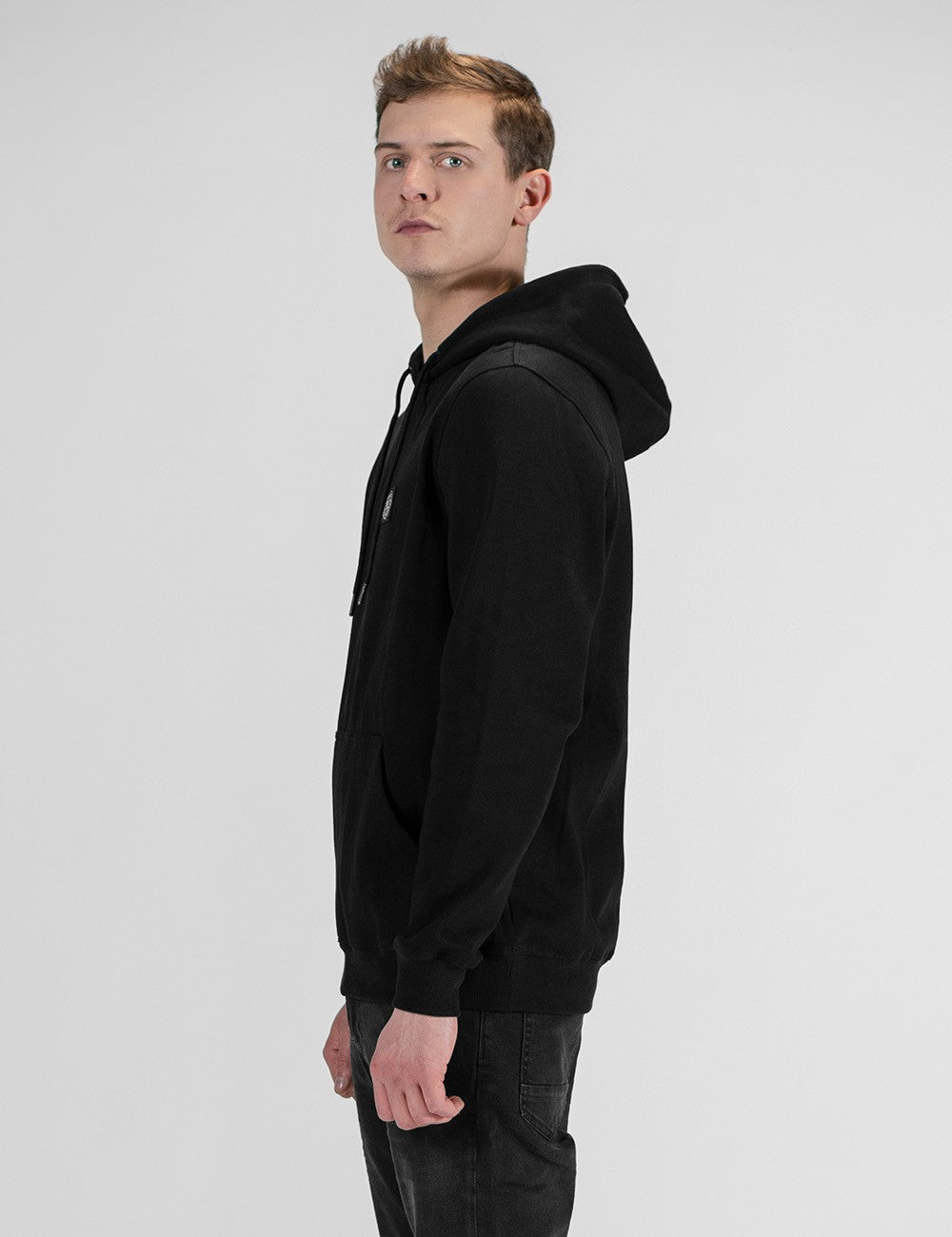 A young man with light brown hair stands in profile against a plain white background, wearing a black drawstring hoodie and dark jeans.