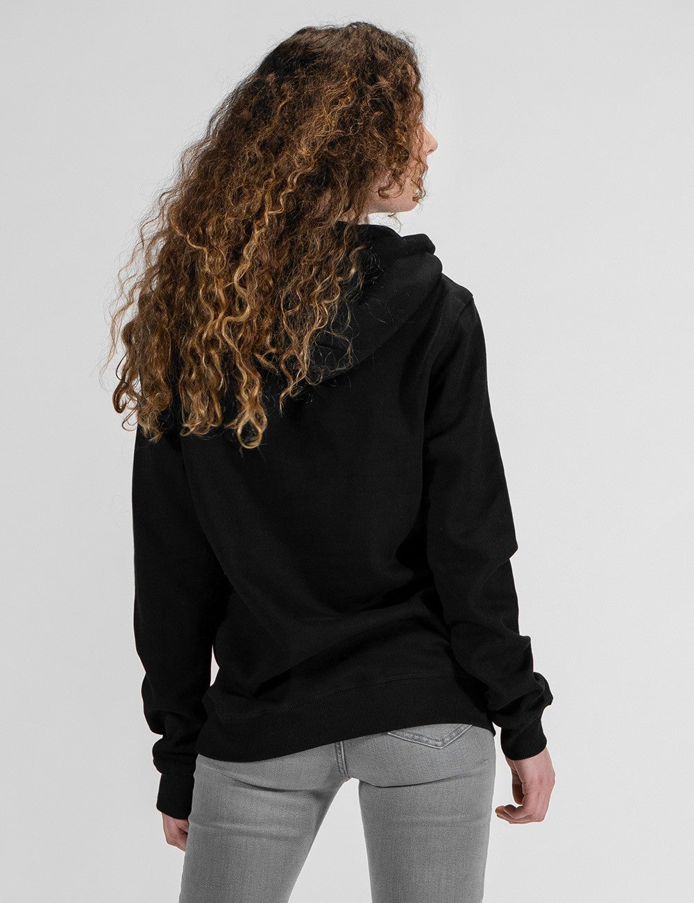 A woman with long, curly blonde and brown hair is captured from behind, showcasing a black hoodie and light gray jeans against a plain white background.