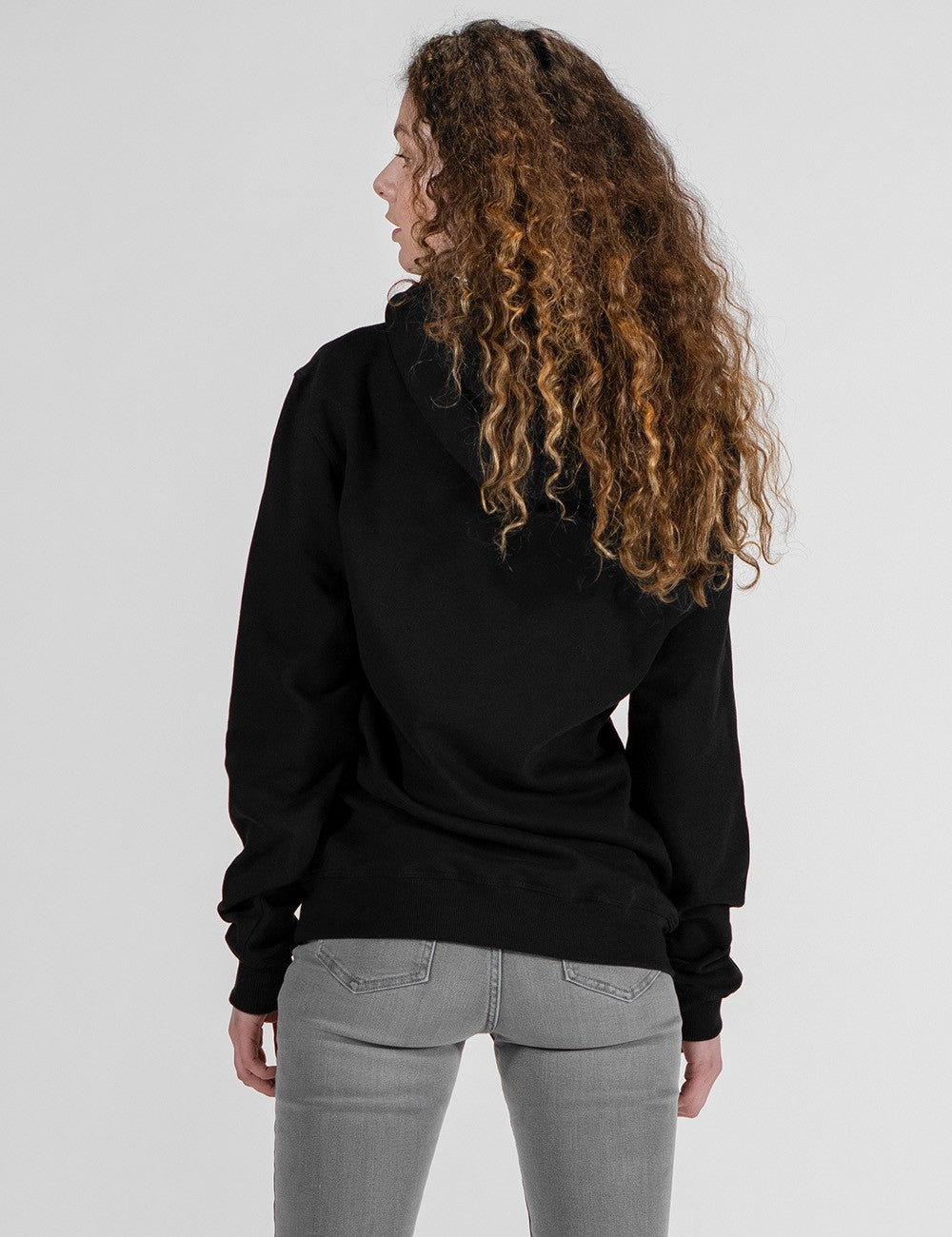 A female model with long, curly, light brown hair is seen from behind, wearing a plain black hoodie and light gray skinny jeans against a solid light gray background.