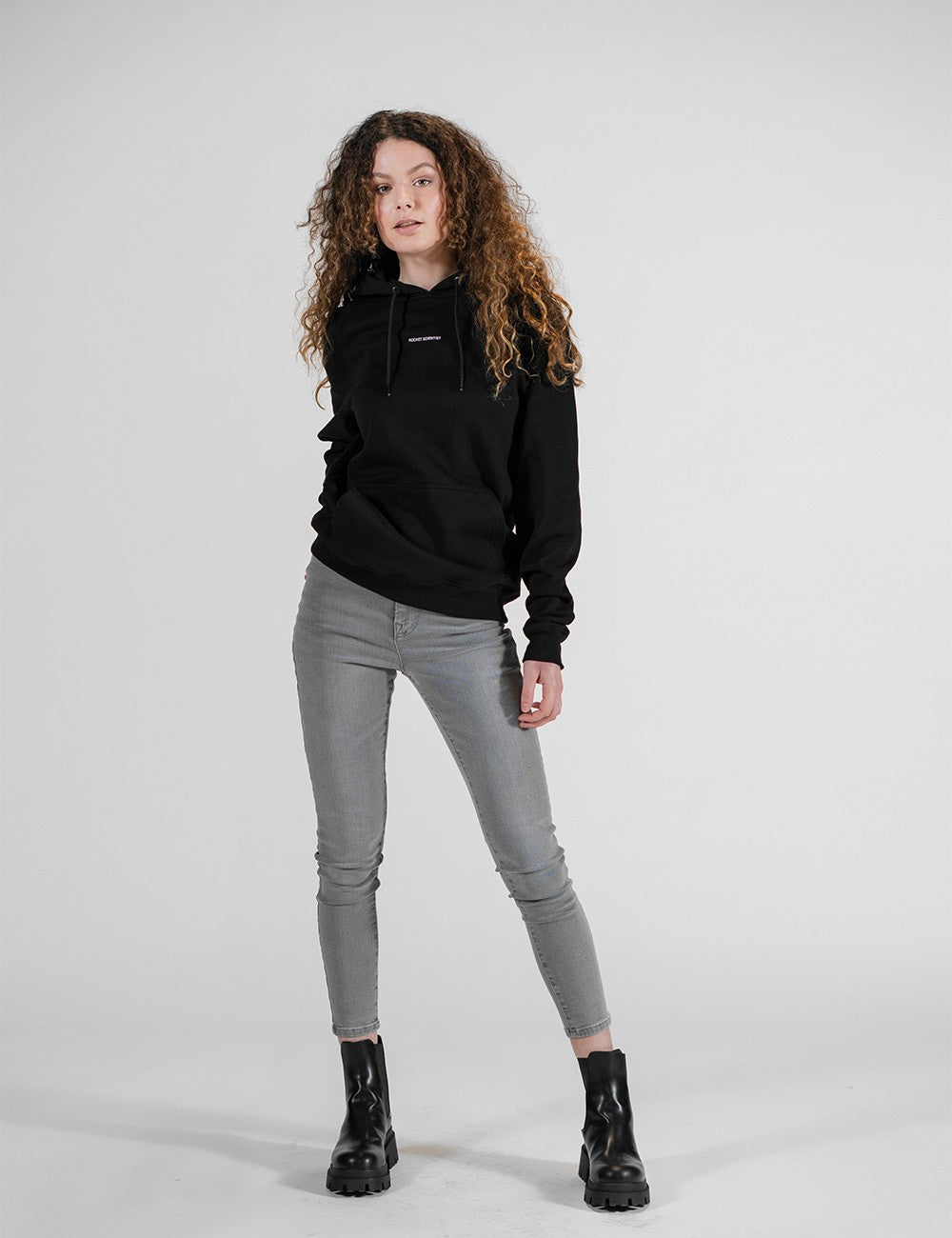 A young model with voluminous, long curly brown hair stands against a plain white background, modeling a casual streetwear outfit. She is wearing a fitted black hoodie with small white text printed across the center chest, slim-fit grey skinny jeans, and chunky black leather combat boots with thick lugged soles. 