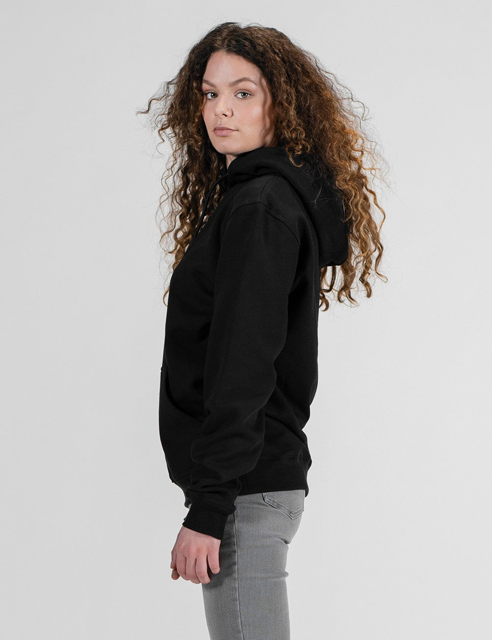 A profile shot of a young woman with voluminous, long curly brown hair looking over her shoulder. She is wearing a plain black hoodie and grey jeans, set against a solid light grey background.