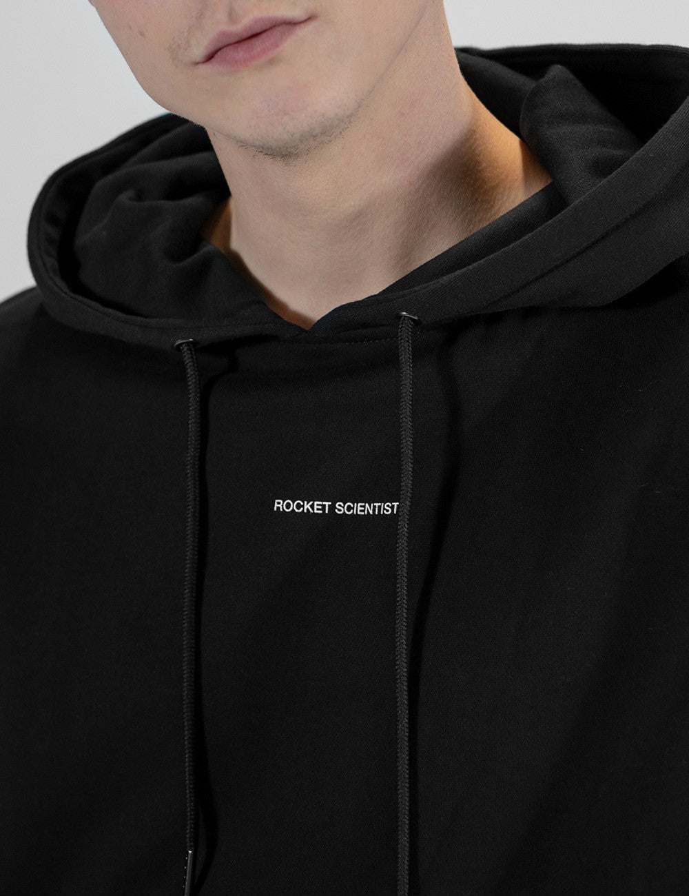 A close-up shot of a person wearing a black hoodie featuring a small, white minimalist text that reads "ROCKET SCIENTIST" centered across the chest. The hood is relaxed around the neck, and two thin black drawstrings hang down over the front of the sweatshirt.