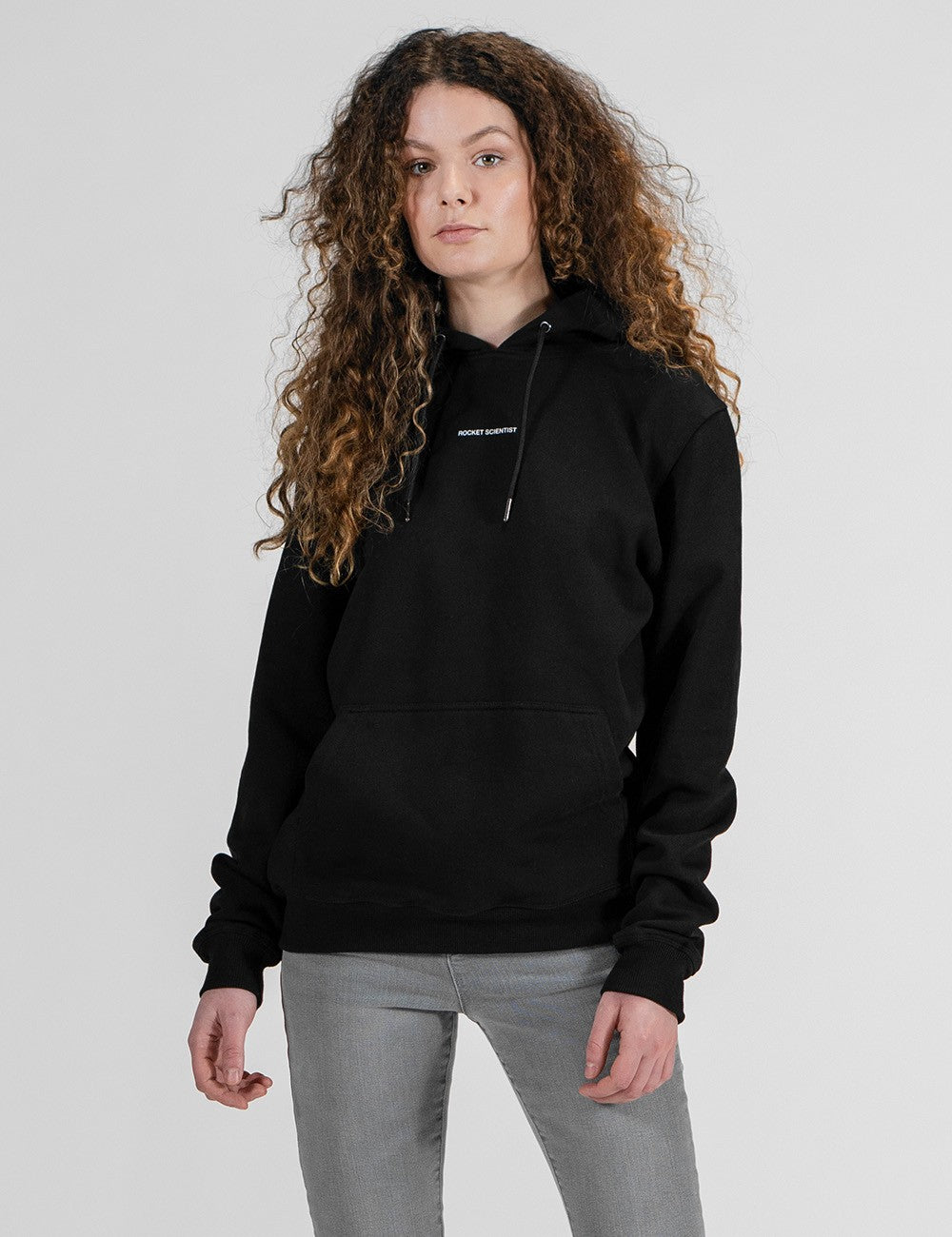 A front shot of a young woman with voluminous, long curly brown hair looking over her shoulder. She is wearing a plain black hoodie with a Rocket Scientist small print in the chest and grey jeans.