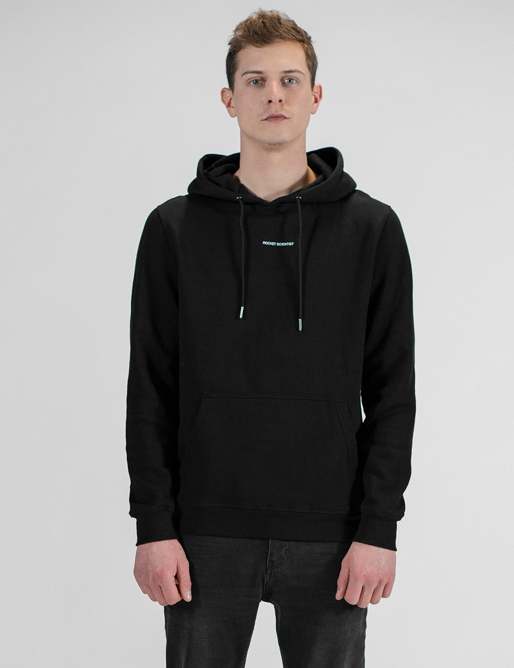 A front profile shot of a man wearing a plain black hoodie and dark jeans, standing against a clean, light grey background.
