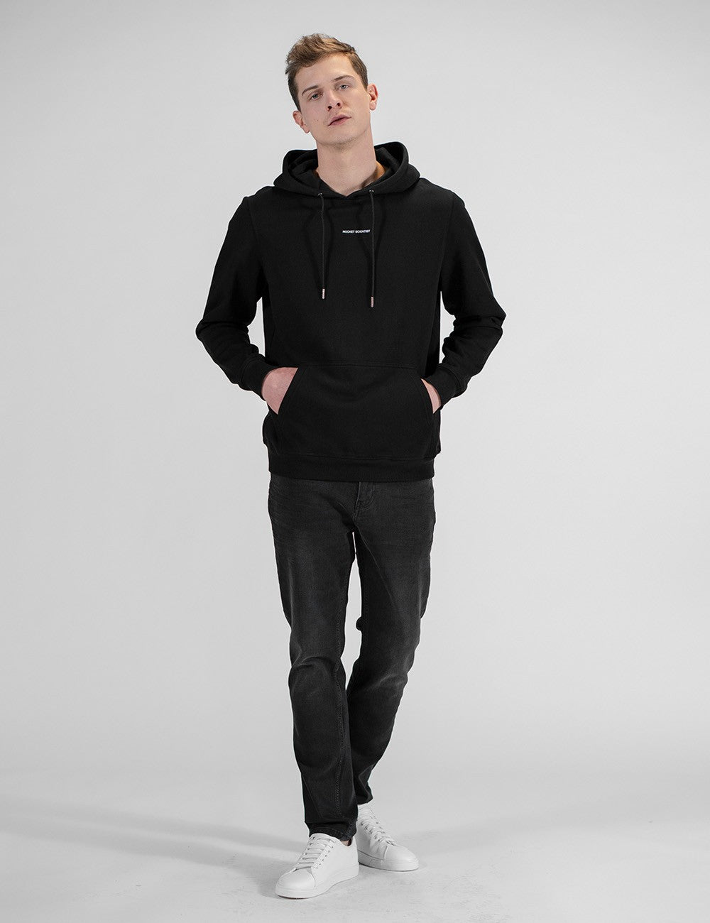 A full-length studio shot of a young man with blonde hair walking forward against a solid light gray background. He is wearing a plain black pullover hoodie with his hands resting inside the front kangaroo pocket. He coordinates the look with dark gray slim-fit jeans and clean white sneakers.
