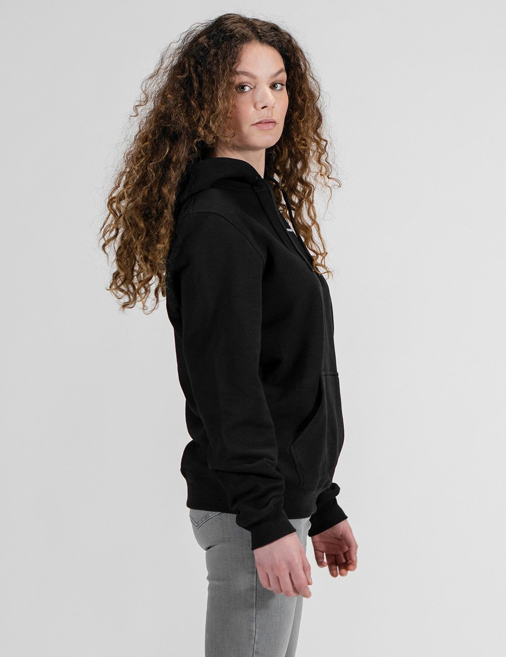 A young woman with long, voluminous, curly brown hair stands in a side-profile view against a plain light gray background. She is looking slightly back toward the viewer over her shoulder. She is wearing a black hoodie with ribbed cuffs and a front pocket, paired with light gray pants.