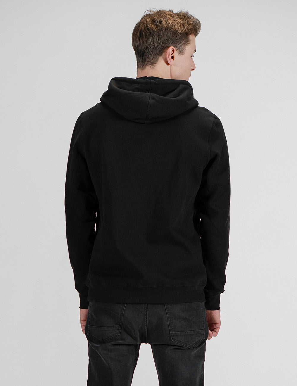A man viewed from behind wears a black pullover hoodie with long sleeves, ribbed cuffs, and a ribbed hem, paired with black pants against a plain gray background.