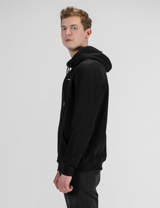 A side profile shot of a man wearing a plain black hoodie and dark jeans, standing against a clean, light grey background.